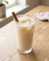 Vaso alto de agua de avena tipo horchata con hielo y una pizca de canela molida, sobre una mesa de madera.